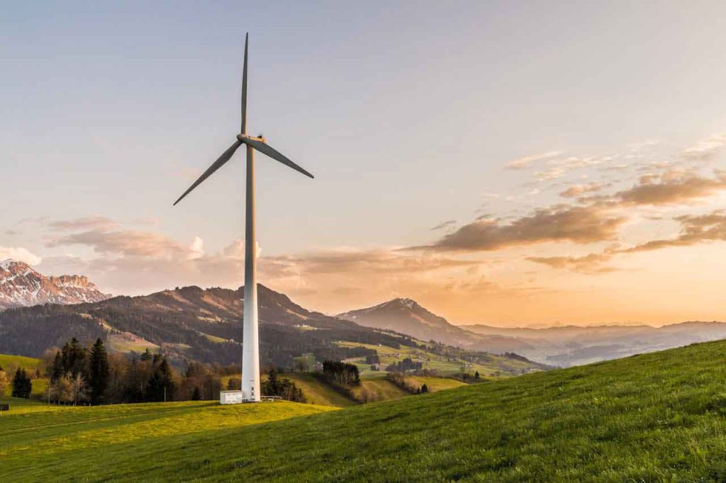Wind turbine on a grassy hillside with mountains in the background at sunset.