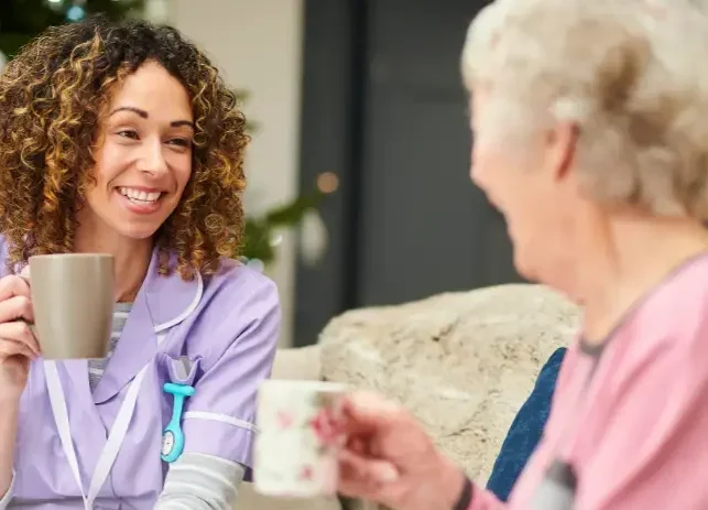 A caregiver and an elderly woman sharing a warm conversation over tea, symbolising companionship and social interaction in care settings.